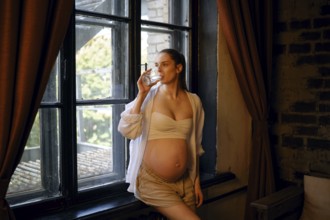 A pregnant woman stands by a large window at home, enjoying a drink from a glass. She wears a
