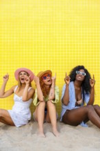 Three diverse women wearing summer clothes and sunglasses are sitting on the sand, pointing upwards