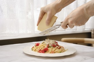 A person grates cheese onto a plate of spaghetti adorned with tomatoes and bacon in a well-lit
