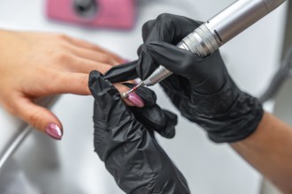 Beautician wearing black gloves is removing nail polish from customer's finger using electric nail