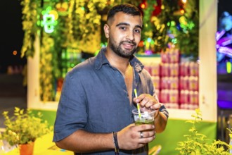 Stylish man savoring a vibrant mojito cocktail at a bustling night market, surrounded by colorful