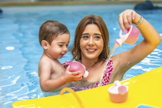 Happy mother and son having fun, playing with pink toys and pouring water in a swimming pool during