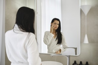 In a bright bathroom, a woman dressed in a white robe applies skincare to her face while looking