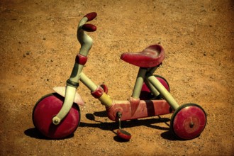 A vintage red tricycle sits abandoned on a sandy ground, showing signs of wear and tear, evoking