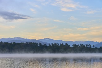 A peaceful lake with a misty surface and mountains in the morning light, Lake Mahinapua, Ruatapu,
