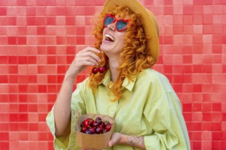 Young redhead woman with heart shaped sunglasses and straw hat is laughing while eating fresh