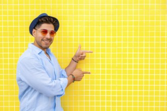 Fashionable man wearing a hat and sunglasses points to the side against a vibrant yellow tiled