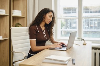 A woman with long curly hair is seated at a stylish wooden desk in a contemporary office. She is
