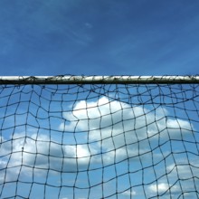 A football goal net stands prominently against a backdrop of a cloudy sky. The bright blue sky adds