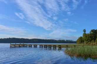 A jetty leads into a calm lake under a blue sky with light clouds, surrounded by lush nature, Lake