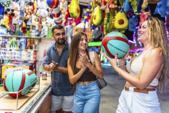 Happy friends enjoying a ball toss game at a lively amusement park, filled with colorful prizes and