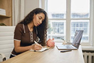 A young woman sits at a wooden desk, diligently writing in her notebook while referring to her