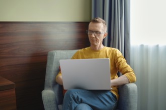 Middle-aged man in a yellow shirt sits comfortably in a grey armchair, focused on his laptop.