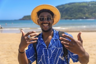 Young man wearing a hat and sunglasses, joyfully gesturing with his hands while enjoying the sunny