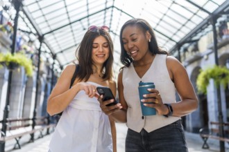Two happy businesswomen walking through a city gallery, sipping coffee and using a smartphone,