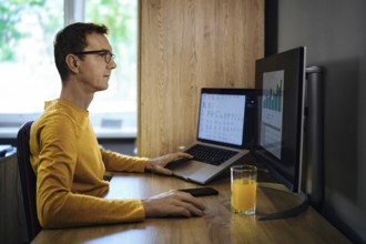 A man wearing glasses sits at a wooden desk in a home office, intently working on a laptop and