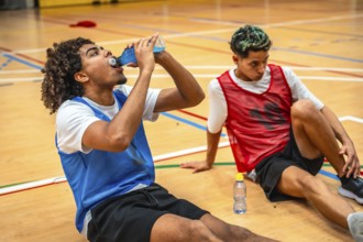 Two basketball players are sitting on the court, taking a break and drinking energy drinks after an