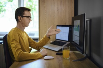 A middle-aged man in a yellow shirt gestures with his hand while presenting data on a monitor in a