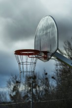 A solitary basketball hoop stands under overcast skies as dusk approaches. The chain net hangs