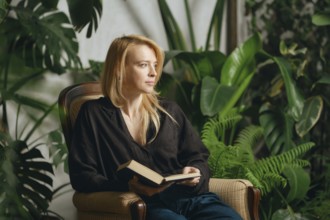 A woman sits comfortably in an armchair holding a book, surrounded by vibrant green plants.She is