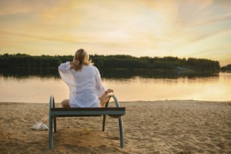 A woman sits peacefully on a bench near the riverbank, enjoying the serene sunset. The soft golden