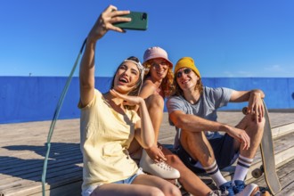 Three cheerful young friends are sitting on a wooden walkway by the sea, taking a selfie with a