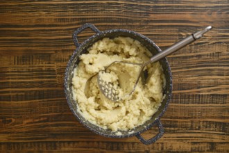 Creamy mashed potatoes are freshly prepared in a pot, sitting on a rustic wooden table. A potato