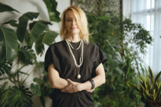 A woman stands gracefully amidst lush greenery in a cozy indoor garden. Her stylish outfit is