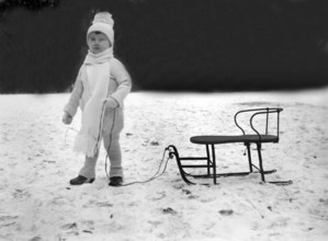 Child with sledge, 1930s, Child in winter clothes with cap and scarf standing next to a sledge in