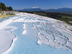 Wide view over white cascade terraces with turquoise blue water against a mountain backdrop, aerial