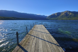 A narrow footbridge leads into the blue waters of a lake, surrounded by an impressive mountain