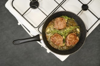 Three thick beef steaks are searing in a skillet on a gas stove. Fresh herbs and garlic surround