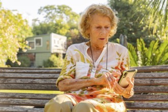 Senior woman sitting on a wooden bench in a sunny park, using a mobile phone to stay connected