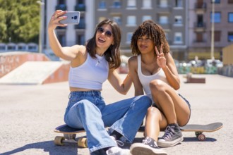 Two young women are taking a selfie with a smartphone while sitting on their skateboards in a skate