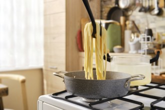A person lifts cooked spaghetti from a pot using a utensil, with steam rising in a cozy kitchen.