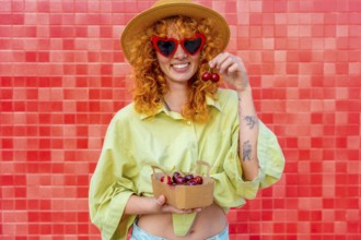 Happy young woman with curly red hair eating cherries from a small cardboard box, wearing heart