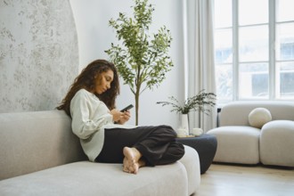 A woman sits comfortably on a light-colored sofa, engaged with her smartphone. She is in a spacious