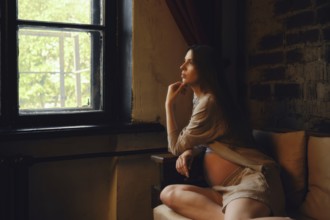 A pregnant woman sits on a couch in her home, looking thoughtfully out of a window. The afternoon