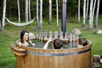 Two children and an their mom enjoy a warm soak in an outdoor wooden hot tub. Lush green grass and