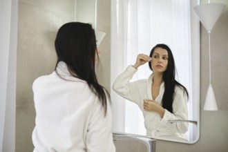 A woman dressed in a white robe stands before a mirror, carefully applying moisturizing serum in a