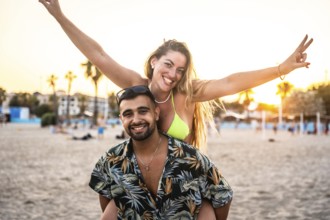 Young couple enjoying piggyback ride on beach, woman making victory sign, sunset in background