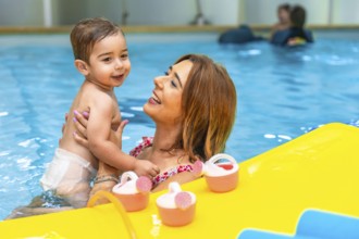Mother holding her smiling son in a swimming pool next to floating toys, enjoying a playful moment