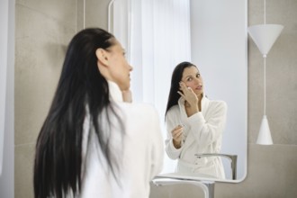 In a stylish bathroom, a woman in a white robe washing off makeup in front of the mirror. She