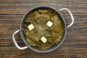 A pot filled with grape leaves prepares to cook dolma on a wooden table. Butter sits on top, ready
