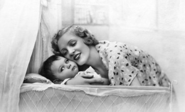 Mother embracing child, 1920s, Smiling mother bends over a baby in a cradle, surrounded by light