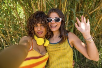 Two happy young women with headphones and sunglasses are taking a selfie and waving in a bamboo