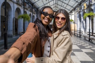 Two joyful women smiling and capturing a selfie beneath a stunning glass and metal roof,