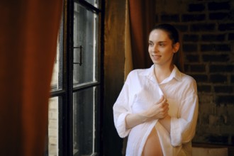 A pregnant woman stands by a large window in her home, wearing a loose white shirt. She smiles