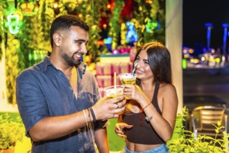 Young man and woman smiling and toasting with cocktails in a vibrant night market setting,