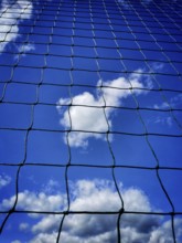 Looking up at a blue sky filled with clouds, framed by a black net. The sunlight highlights the